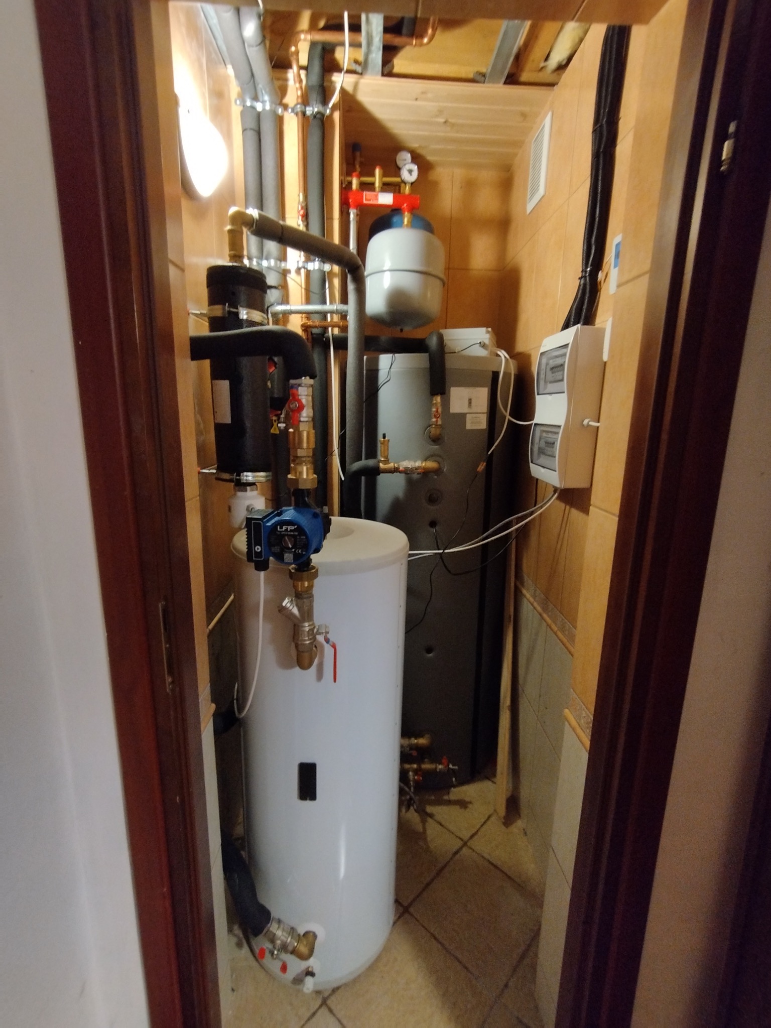Zestaw kotłowni w małym pomieszczeniu z białym zasobnikiem ciepłej wody, szarym kotłem i instalacją rurową. Widoczne zabezpieczenia i osprzęt.