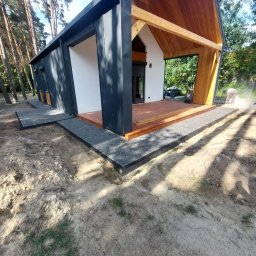 STYL-REMONT - Nowoczesny dom z drewnianym tarasem i ścieżką z szarej kostki. W tle las, a w kadrze widoczny pracownik podczas prac wykończeniowych.