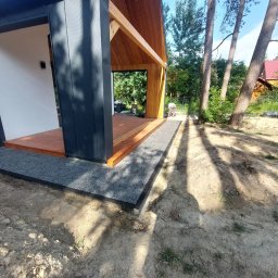 STYL-REMONT - Nowoczesny taras z drewnianą podłogą i obrzeżem z drobnego kamienia, otoczony zielenią i drzewami. Widoczna część konstrukcji budynku z drewnianymi elementami.