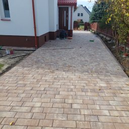 STYL-REMONT - Ułożona kostka brukowa w odcieniach beżu i brązu tworzy wzór na podjeździe. Widoczny fragment domu z czerwonym dachem i pracownik w trakcie pracy.