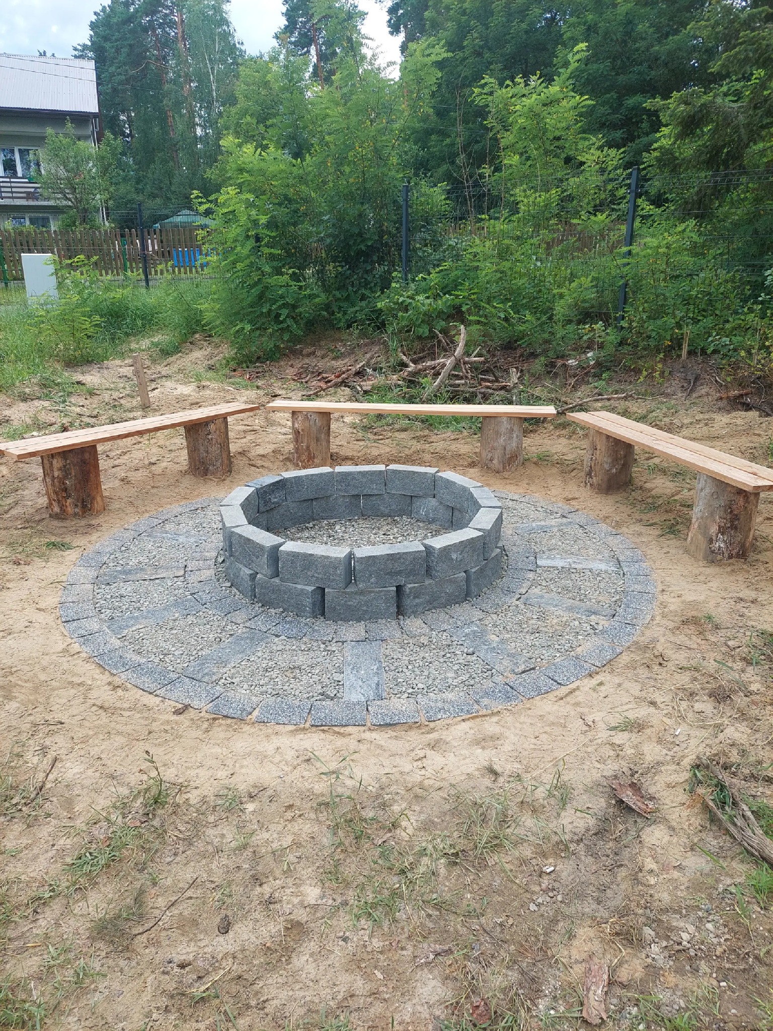 Ognisko z szarej kostki brukowej otoczone okręgiem z drobnego żwiru, z drewnianymi ławkami na pniakach w ogrodzie. Widok z góry, naturalne oświetlenie.