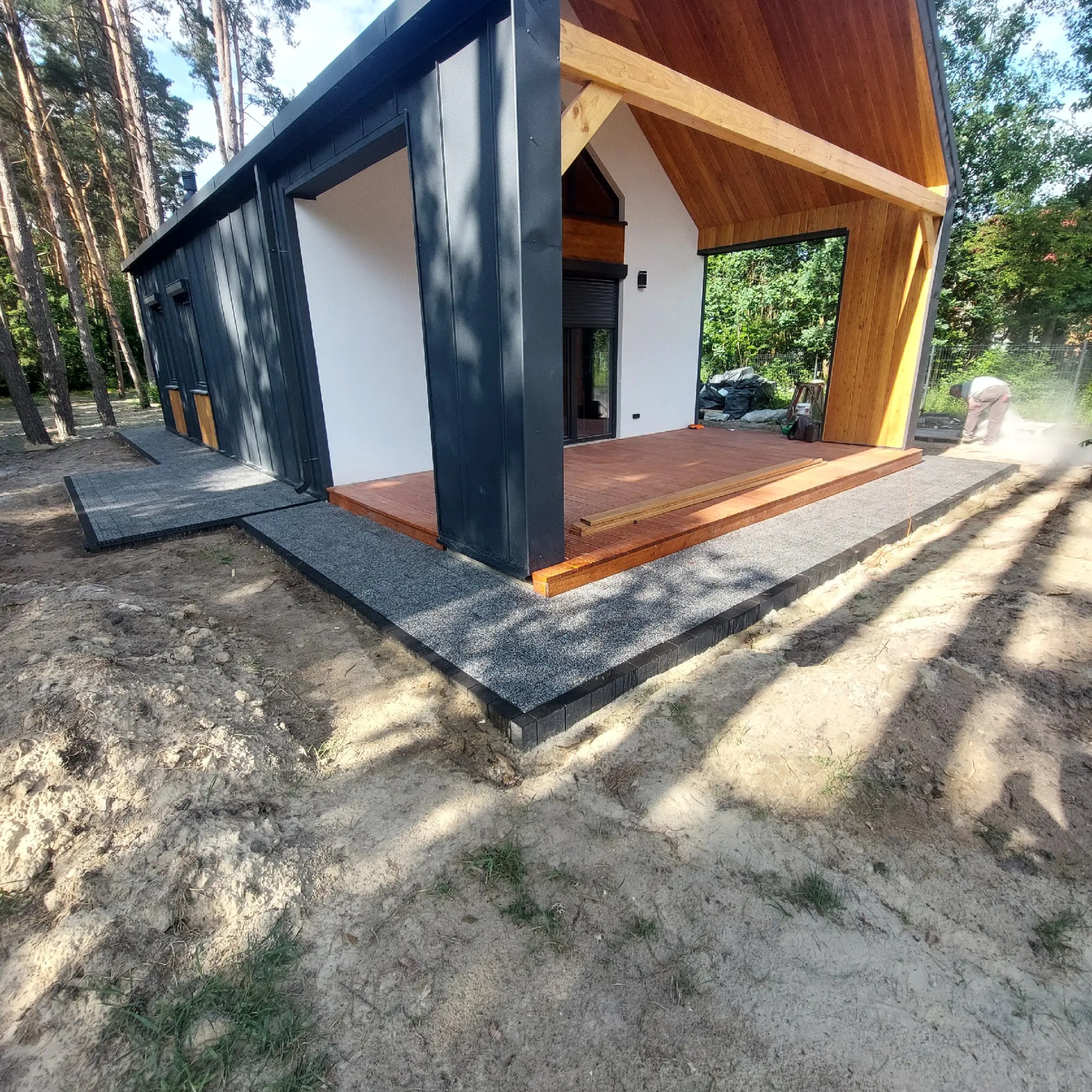 Nowoczesny dom z drewnianym tarasem i ścieżką z szarej kostki. W tle las, a w kadrze widoczny pracownik podczas prac wykończeniowych.