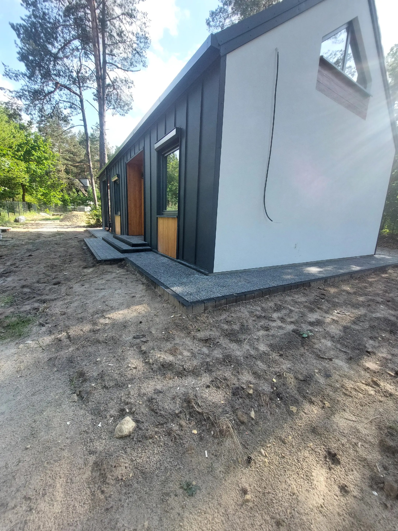 Nowoczesny, parterowy budynek z szarą elewacją i drewnianymi akcentami, otoczony ziemią i zielenią. Wykończone wejście z kostki brukowej.