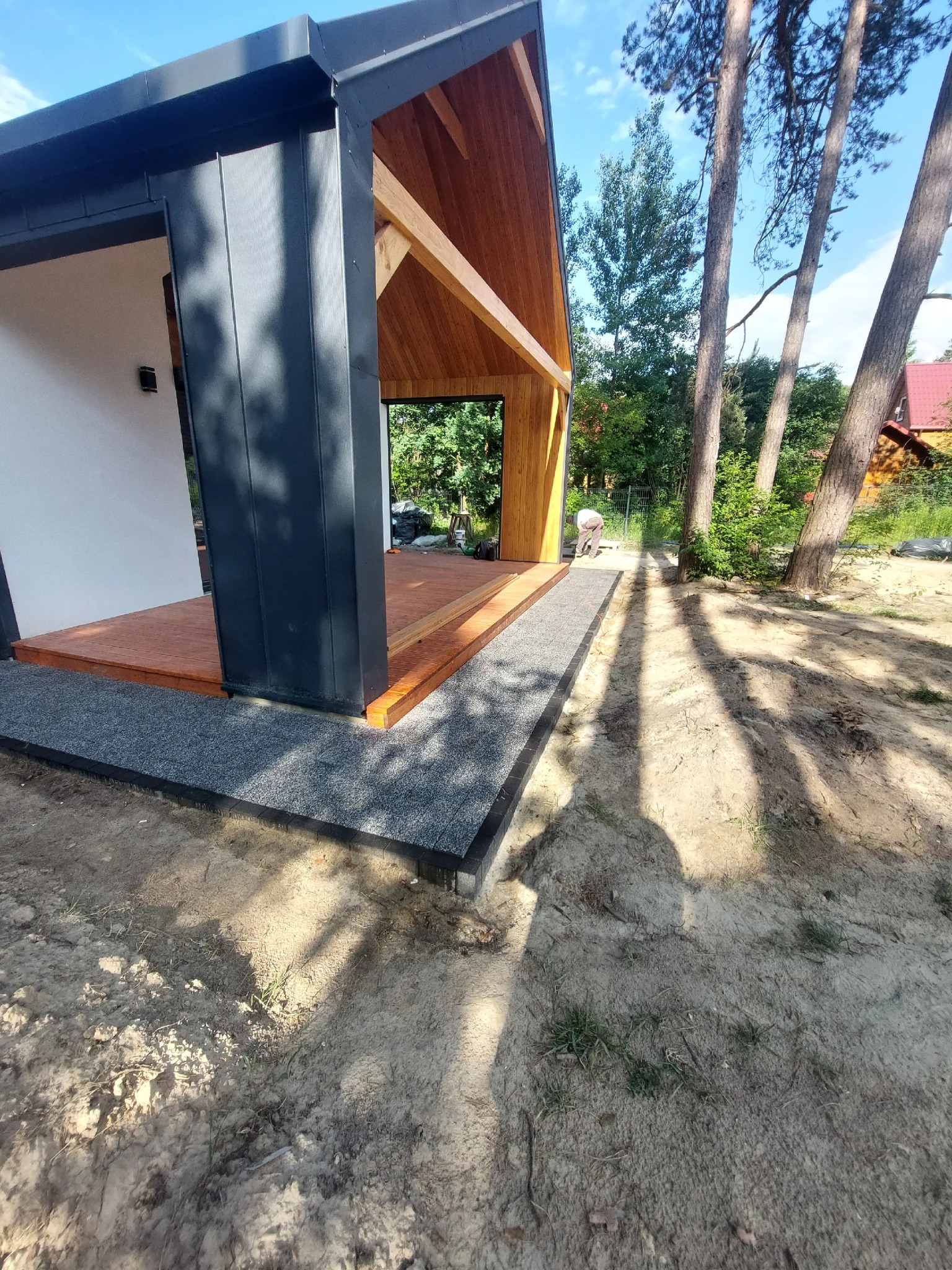 Nowoczesny taras z drewnianą podłogą i obrzeżem z drobnego kamienia, otoczony zielenią i drzewami. Widoczna część konstrukcji budynku z drewnianymi elementami.