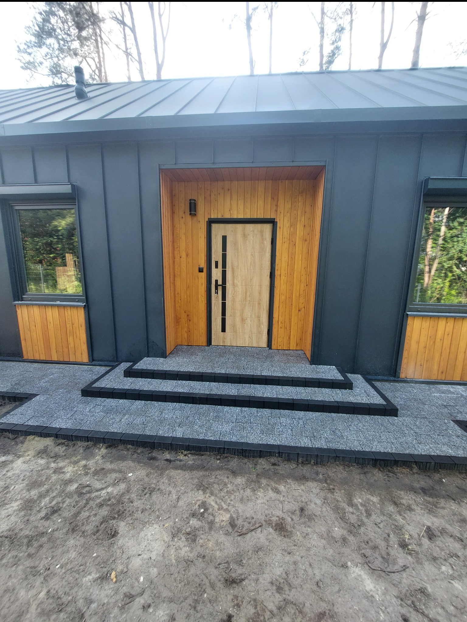 Nowoczesny dom z szarą elewacją, drewnianymi akcentami i brukowanym wejściem z trzema stopniami. Widoczne okna i drzwi wejściowe.