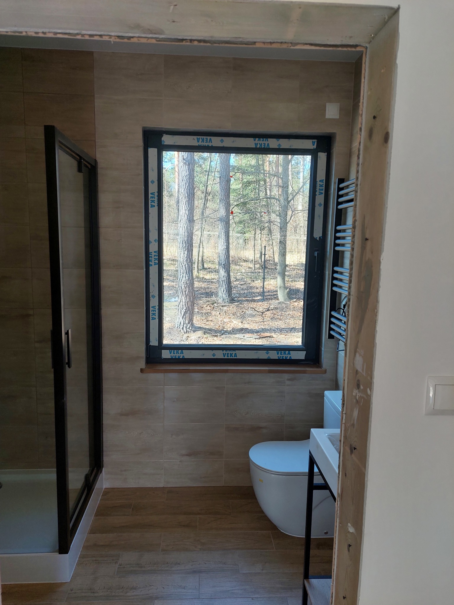 Jasna łazienka z kabiną prysznicową, umywalką i toaletą. Okno z widokiem na las. Płytki imitujące drewno na podłodze i ścianach. Grzejnik drabinkowy obok okna.