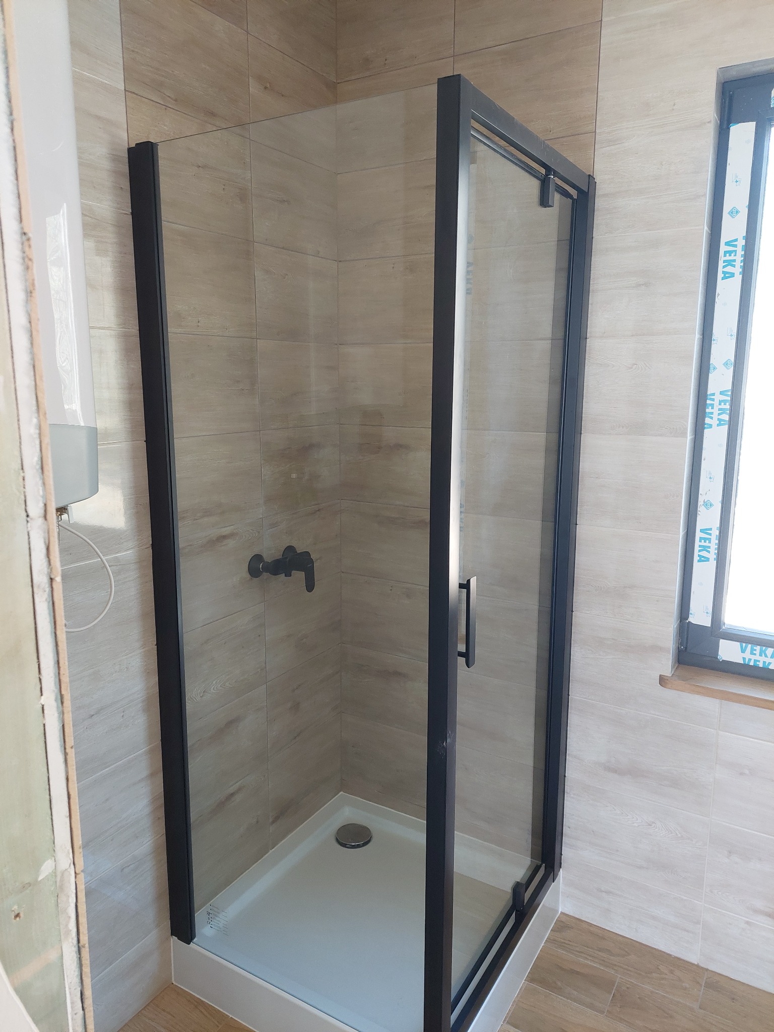 Nowoczesna kabina prysznicowa z czarnymi profilami i beżowymi płytkami imitującymi drewno. Widoczny fragment okna i armatura w tym samym kolorze.