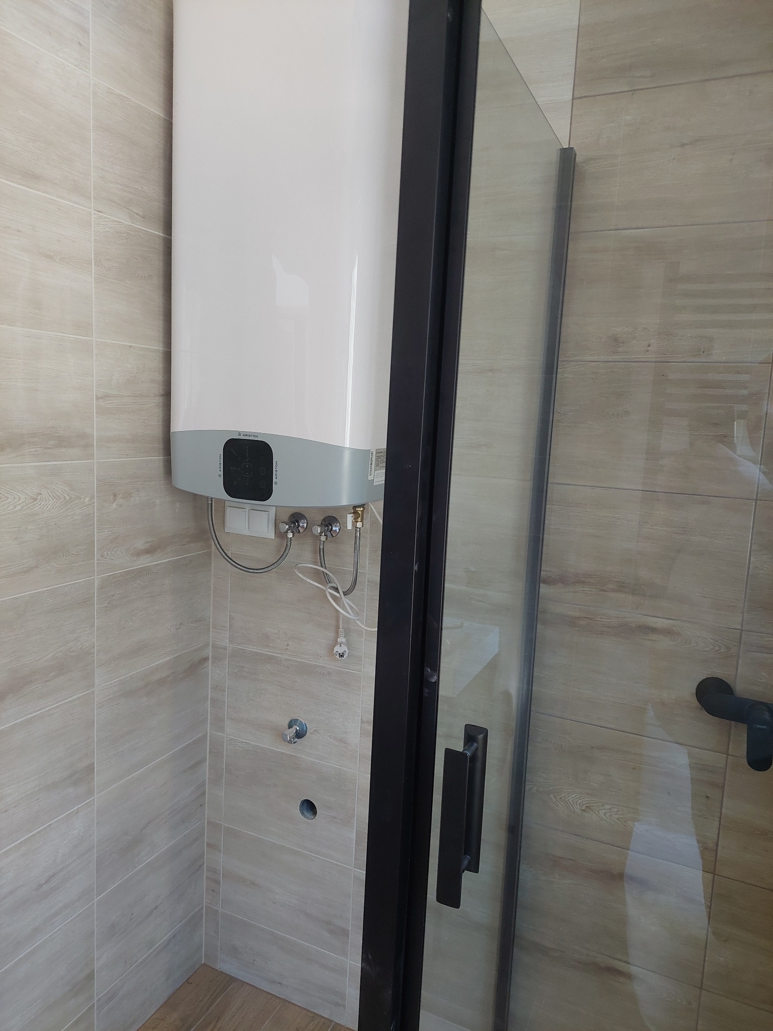 Bojler Ariston zamontowany w łazience z beżowymi płytkami imitującymi drewno, obok kabiny prysznicowej z czarną ramą.