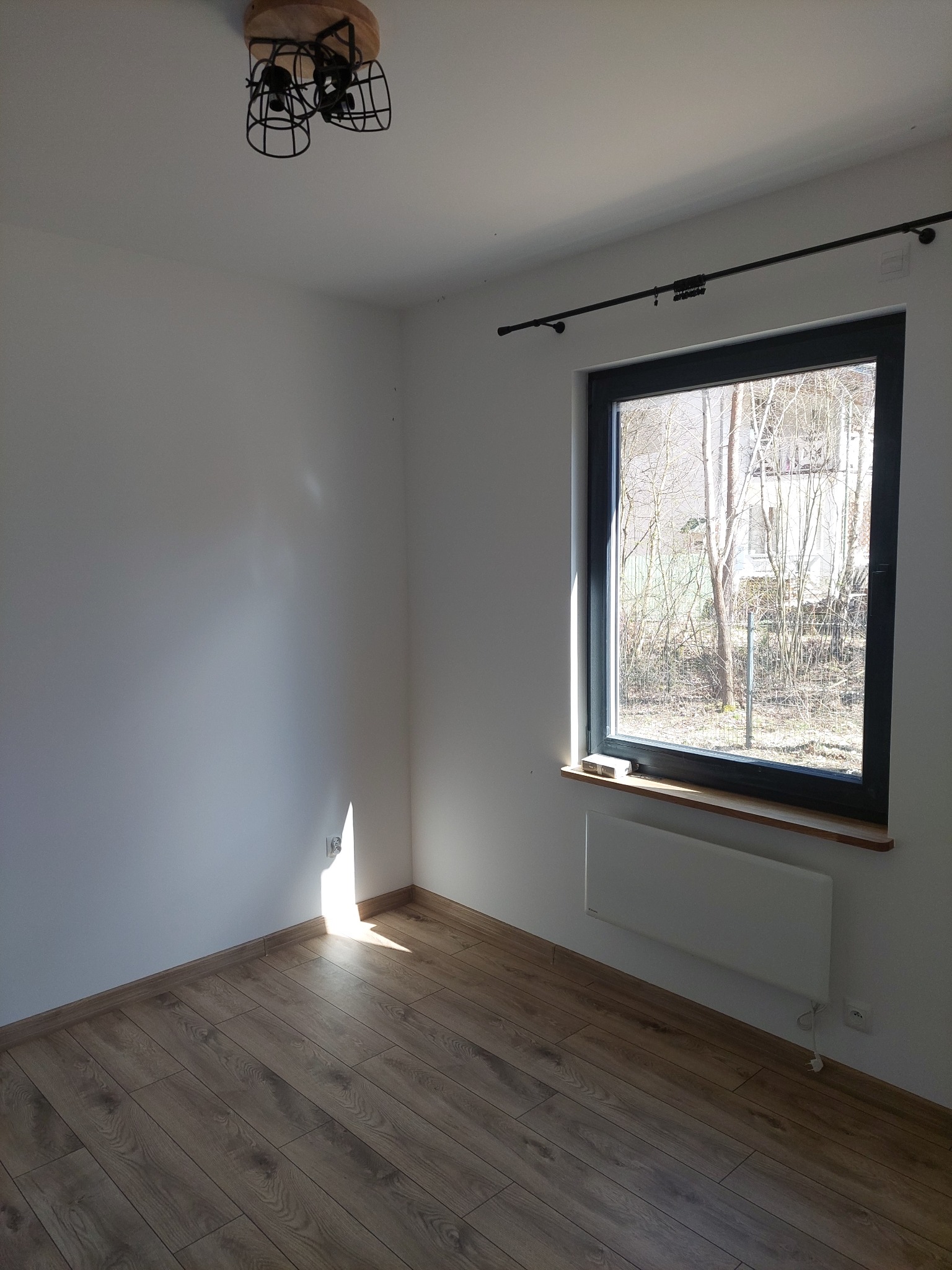 Jasne wnętrze pokoju po remoncie: drewniana podłoga, białe ściany, okno z widokiem na zieleń, nowoczesna lampa sufitowa i grzejnik elektryczny pod oknem.