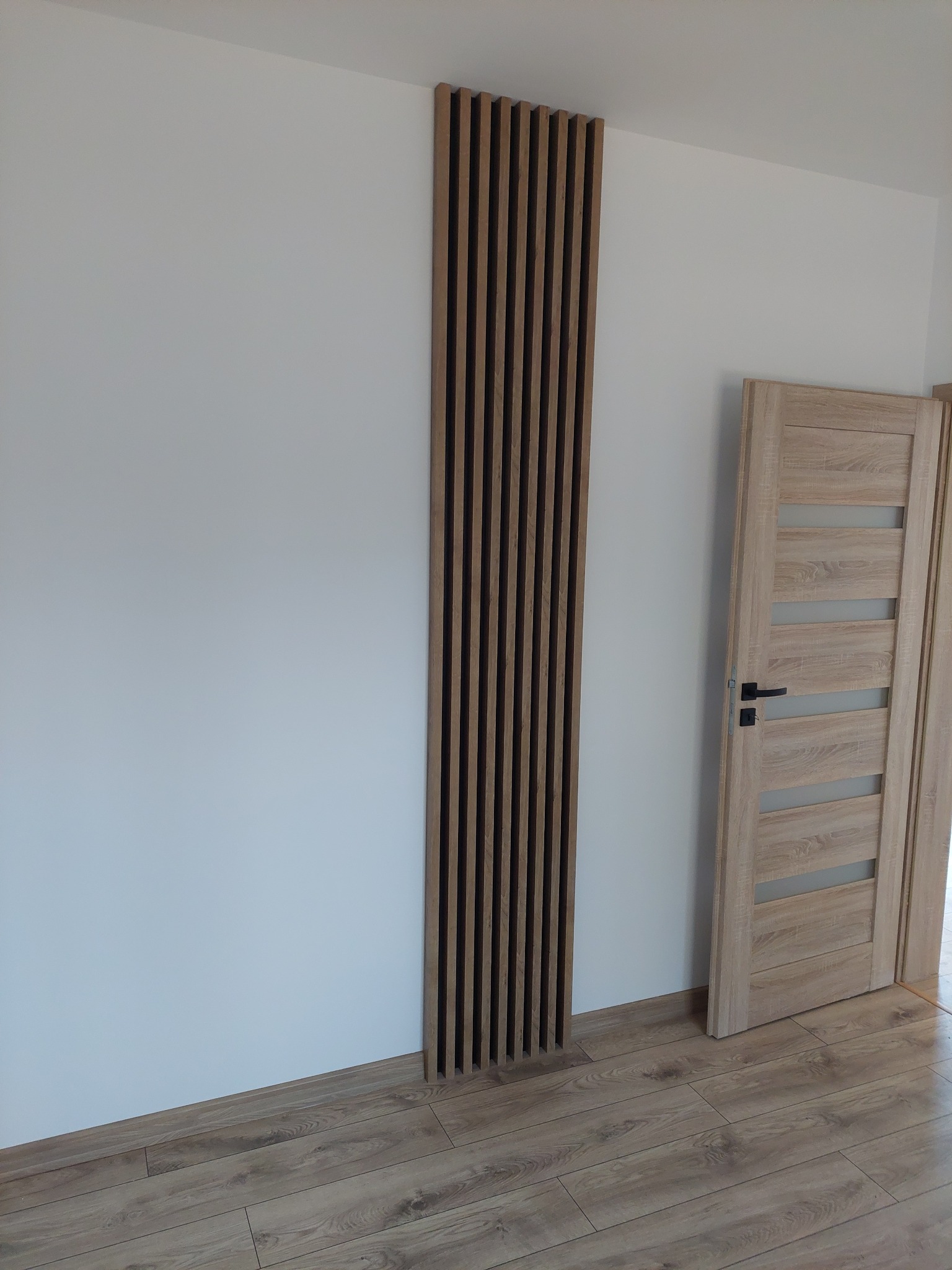 Drewniana lamelowa dekoracja ścienna w pionie, obok drzwi z przeszkleniami, na tle białej ściany i podłogi z paneli w odcieniach brązu. Minimalistyczny design.