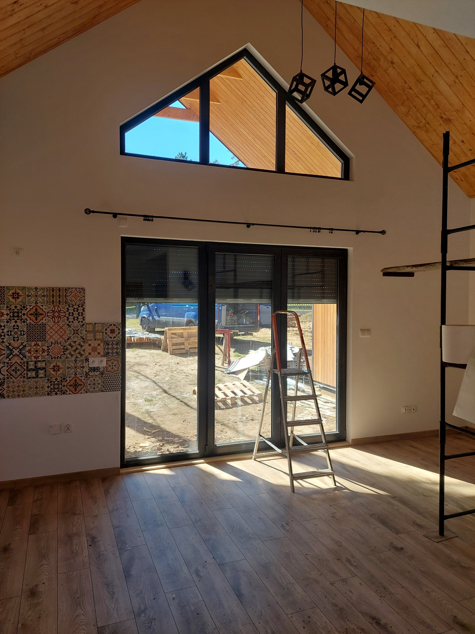 Wnętrze z drewnianą podłogą i dekoracyjnymi płytkami ściennymi. Widoczne okno z roletami, drabina i drewniany sufit. Nowoczesne lampy wiszące.