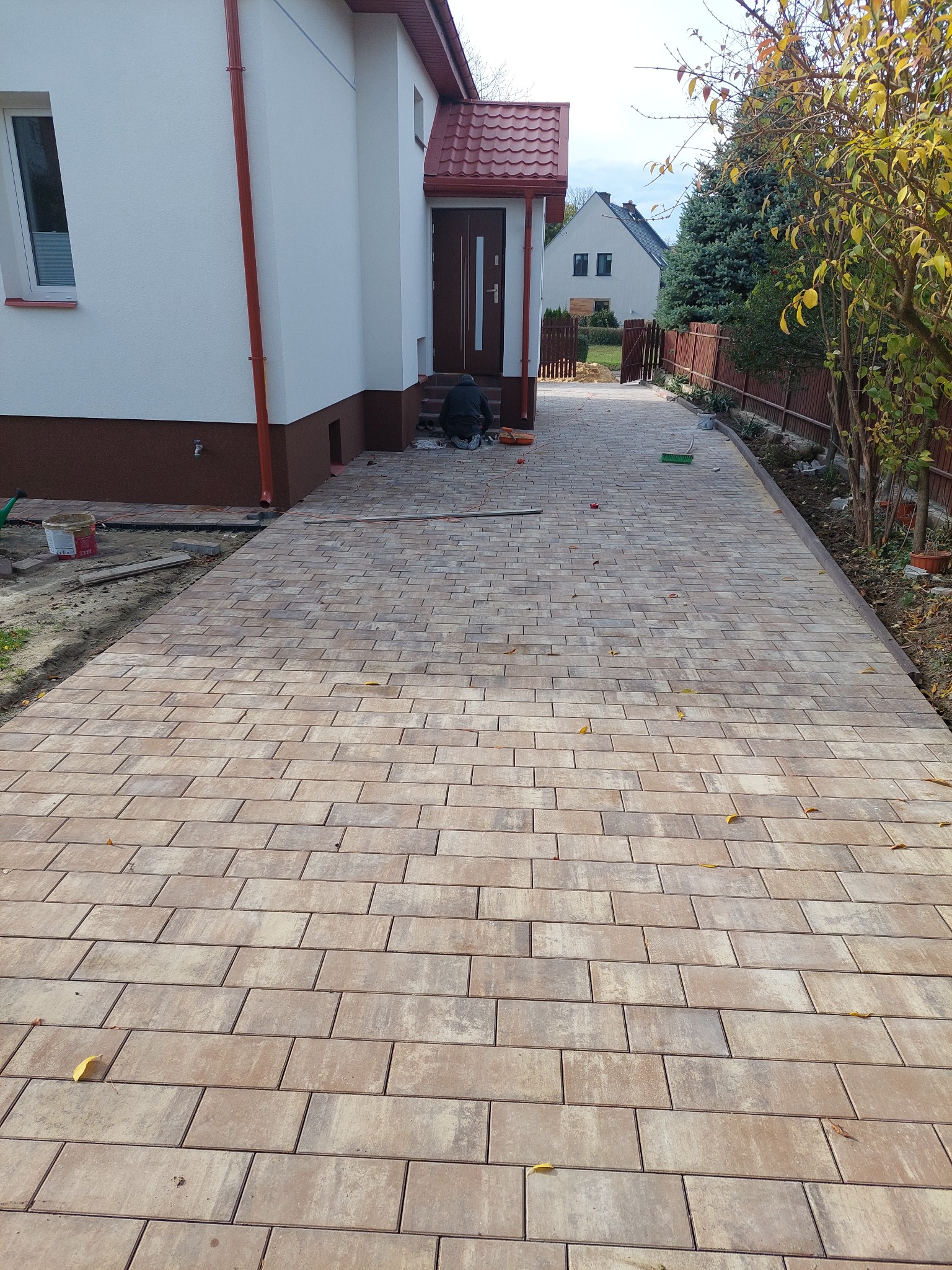 Ułożona kostka brukowa w odcieniach beżu i brązu tworzy wzór na podjeździe. Widoczny fragment domu z czerwonym dachem i pracownik w trakcie pracy.