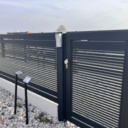 Daniel Zakrzewski Ogrodzenia - Nowoczesna, antracytowa brama z poziomymi lamelami i domofonem, uzupełniona o niski fundament i żwirowe podłoże. Widoczne tabliczki informacyjne.