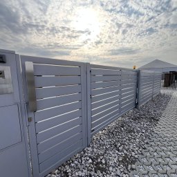 Daniel Zakrzewski Ogrodzenia - Nowoczesna, pozioma brama wjazdowa z szarego metalu z domofonem i skrzynką na listy, na tle brukowanej drogi i pochmurnego nieba.