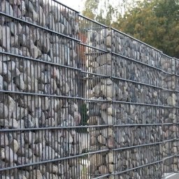 Ogrodzenia Zakrzewski - Gabionowy płot z kamieniami, z metalową siatką, widok z boku na tle zieleni. Nietypowe wypełnienie, poziome linie siatki, regularny układ kamieni.