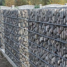 Daniel Zakrzewski Ogrodzenia - Gabionowe ogrodzenie z kamieniami w metalowej siatce, widok z boku, betonowa podstawa, w tle drzewa.