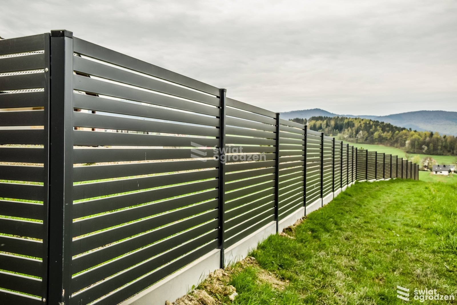 Nowoczesne ogrodzenie panelowe z poziomych, czarnych listew na betonowej podmurówce. Widok na zielony trawnik i zalesione wzgórza w tle.
