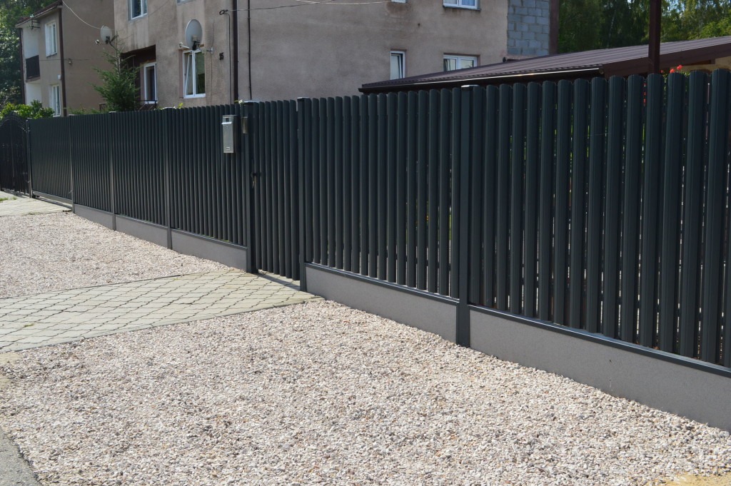 Długi, grafitowy płot ze sztachet z furtką i skrzynką na listy, na tle domu z anteną satelitarną, obok ścieżka z kostki i żwir.