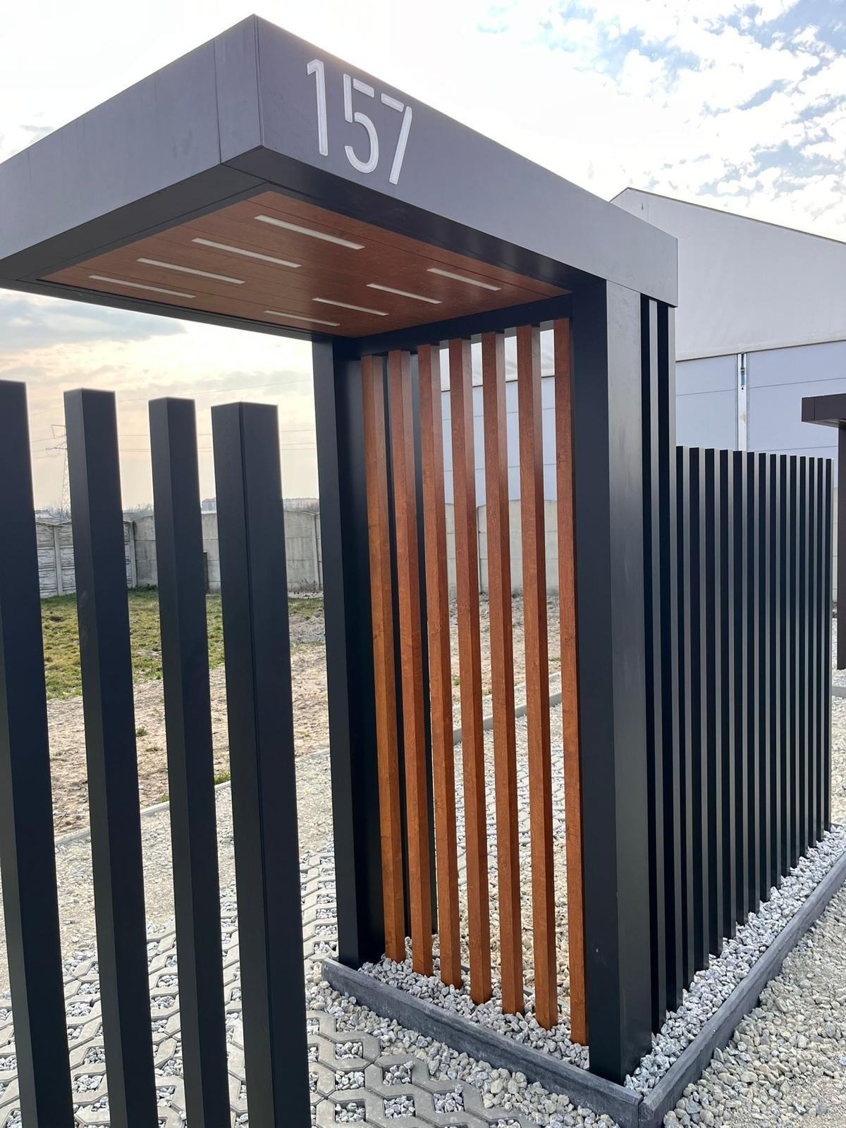 Nowoczesna konstrukcja wejściowa z numerem 157, z elementami drewna i metalu, otoczona kamiennym wykończeniem. Minimalistyczny design.