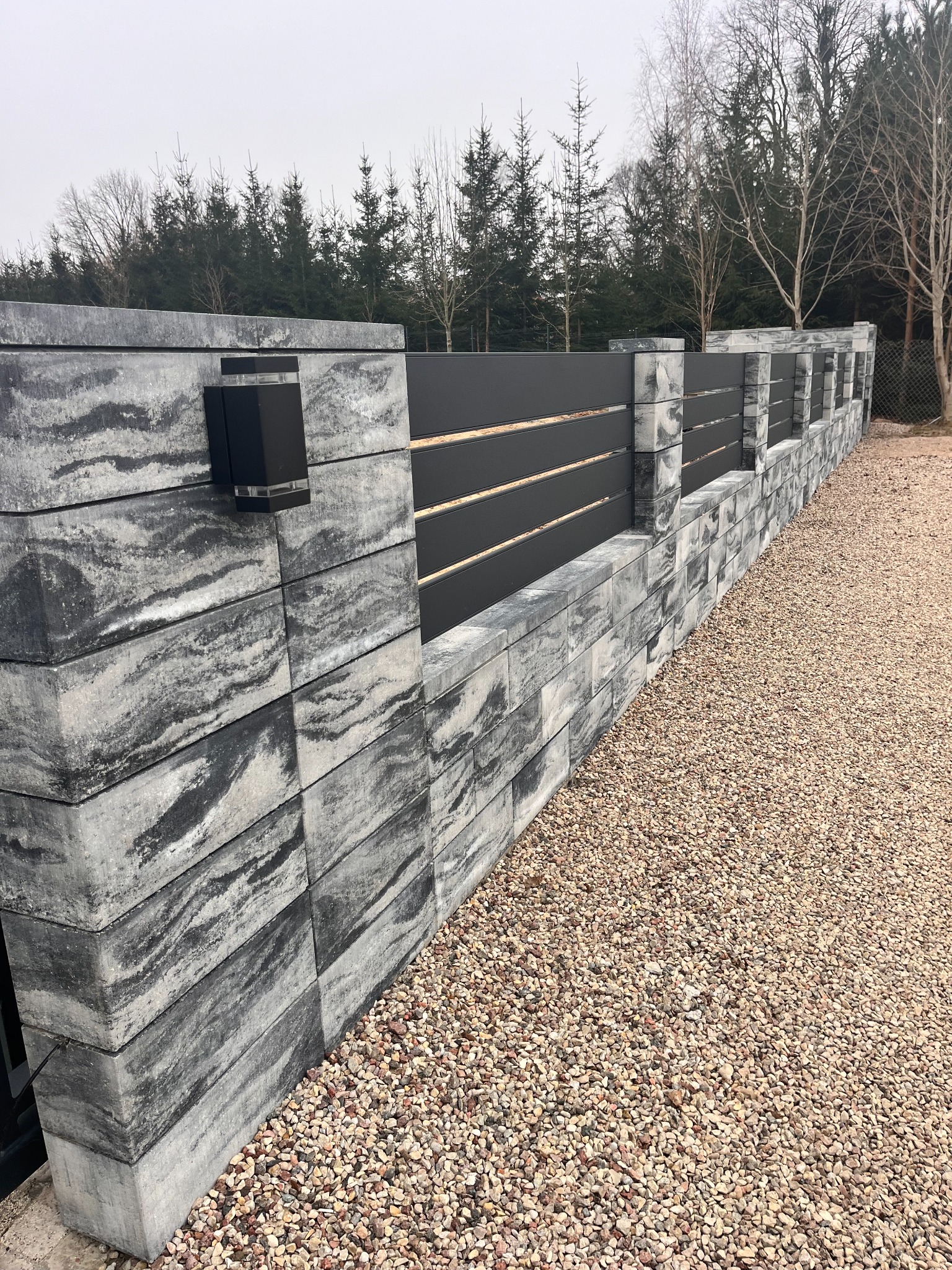 Nowoczesne ogrodzenie z szarego betonu imitującego kamień i czarnych paneli, oświetlone lampami na słupkach, na tle drzew i żwirowego podłoża.