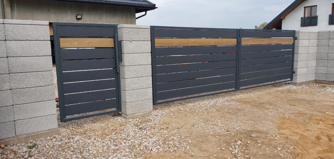 Nowoczesna brama wjazdowa z szarego metalu z poziomymi panelami i drewnianymi akcentami, osadzona na betonowych słupkach z szarej kostki.