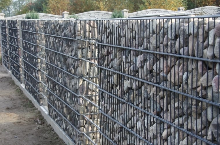 Gabionowe ogrodzenie z kamieniami w metalowej siatce, widok z boku, betonowa podstawa, w tle drzewa.