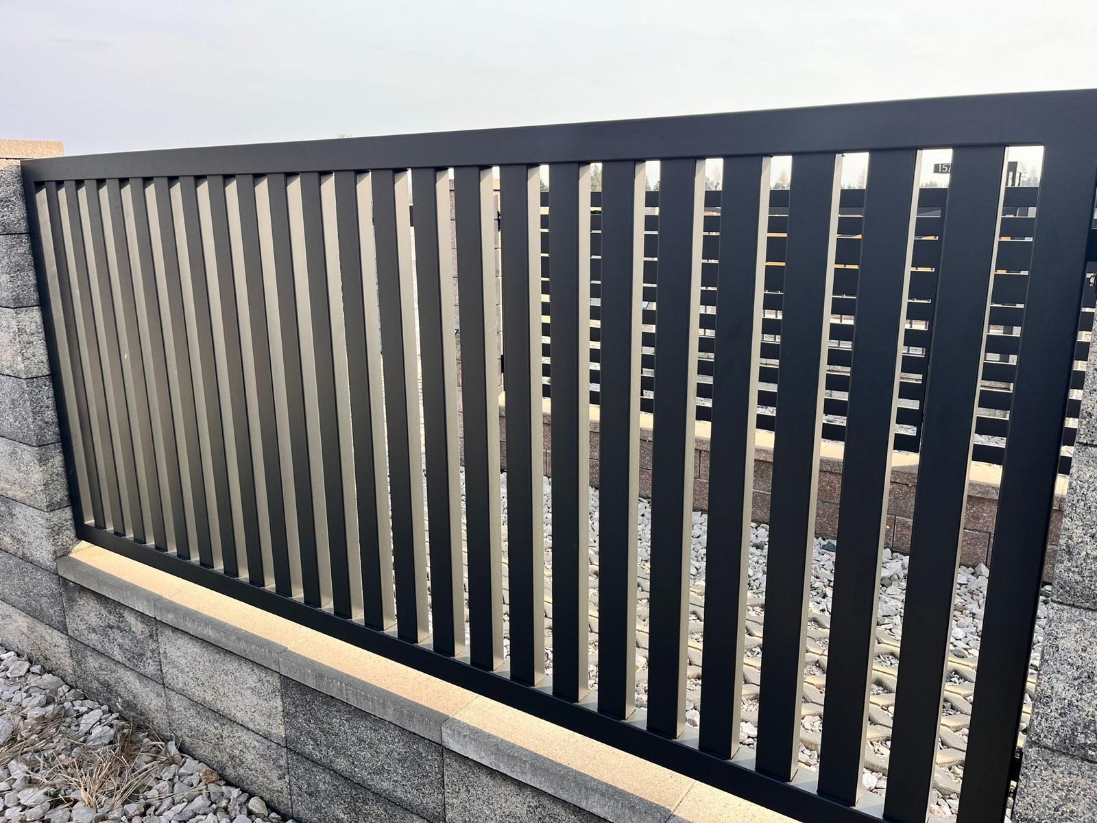 Nowoczesne ogrodzenie panelowe w kolorze grafitowym, z pionowymi szczebelkami, osadzone na niskiej, kamiennej podmurówce. Widoczne detale konstrukcyjne.