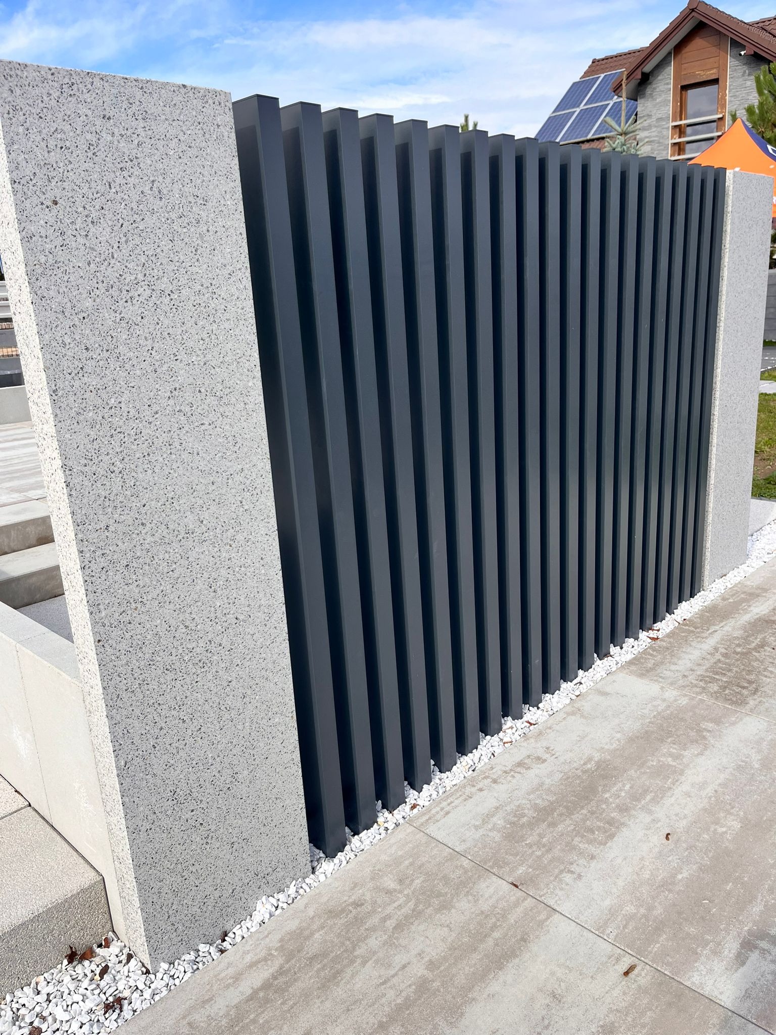 Nowoczesne ogrodzenie: grafitowe, pionowe panele między szarymi, teksturowanymi słupkami. Białe kamyczki u podstawy, dom z panelami słonecznymi w tle.