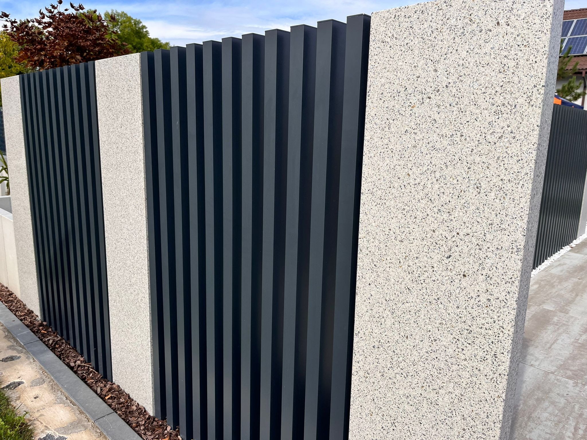 Nowoczesne ogrodzenie z szarych, pionowych paneli i białych, teksturowanych słupków. Widoczne detale wykończenia i fragment otoczenia.