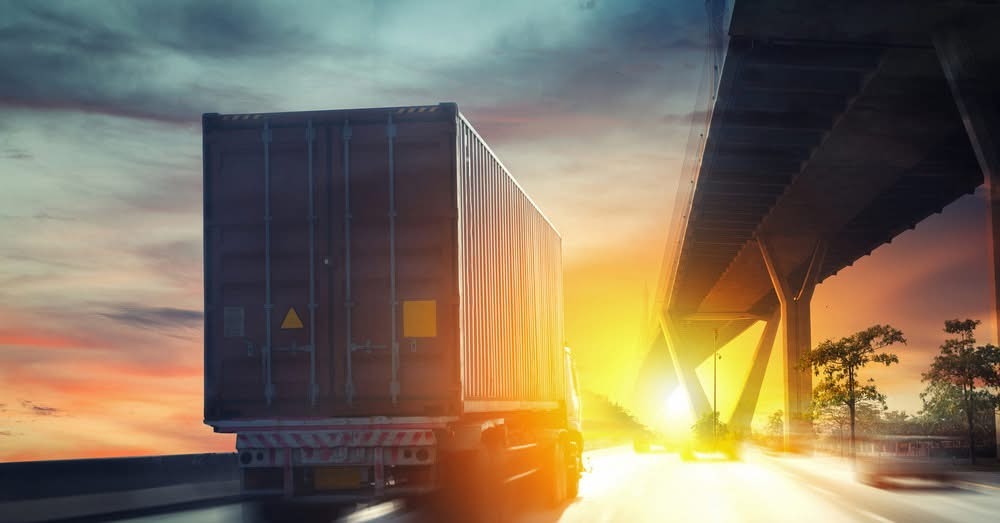 Ciężarówka z kontenerem w ruchu pod wiaduktem, pomarańczowe niebo o zachodzie słońca, dynamiczne ujęcie z efektem rozmycia ruchu.