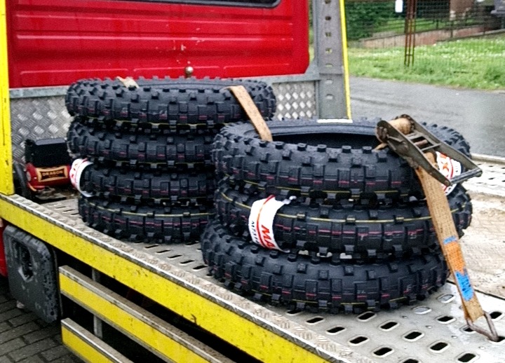 Stosy opon motocyklowych z agresywnym bieżnikiem, zabezpieczone pasami transportowymi na platformie lawety. Widoczne detale opon i mocowanie.