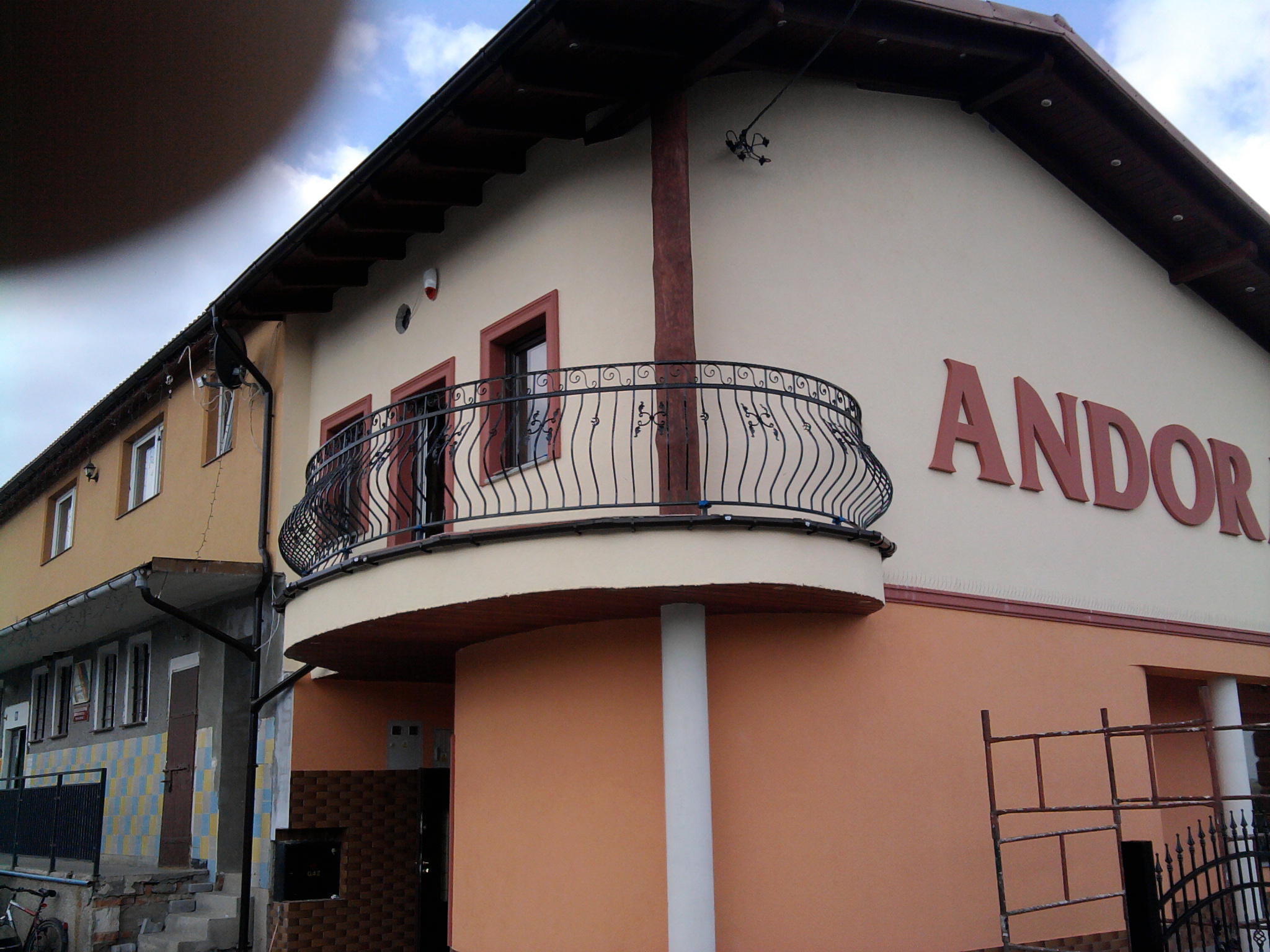 Balkon z metalową, ozdobną balustradą przy budynku z napisem 'ANDOR' na elewacji, widoczne rusztowanie.