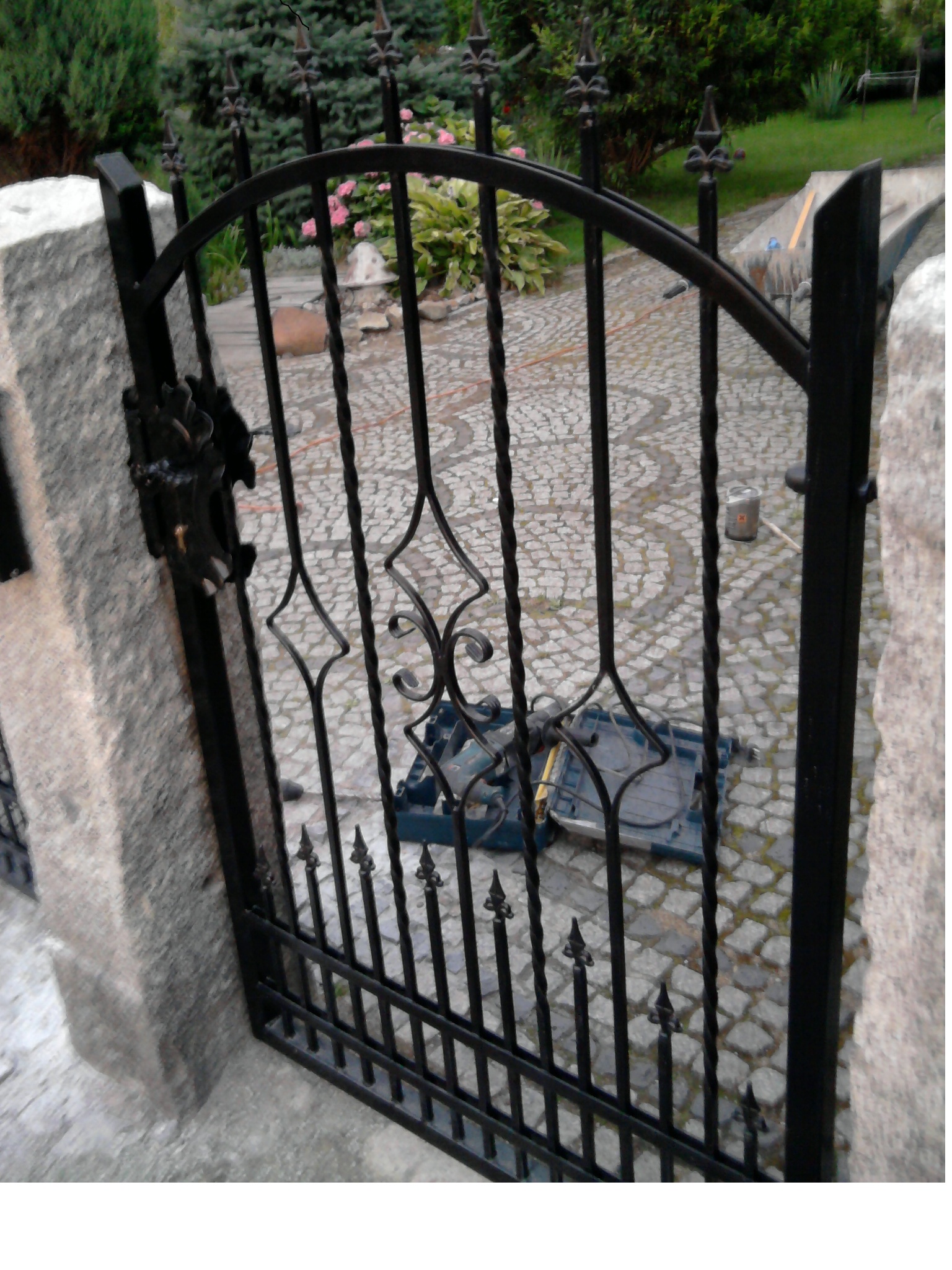 Kuta, czarna furtka z metaloplastyki zamontowana między kamiennymi słupkami, z widoczną skrzynką narzędziową na brukowanym podłożu.