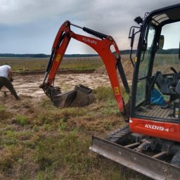 Usługi mini koparką Bogusław Kureń - Pomarańczowa mini koparka Kubota KX019-4 w trakcie prac ziemnych na zewnątrz, z operatorem w tle i wykopem w oddali. Pochmurne niebo.