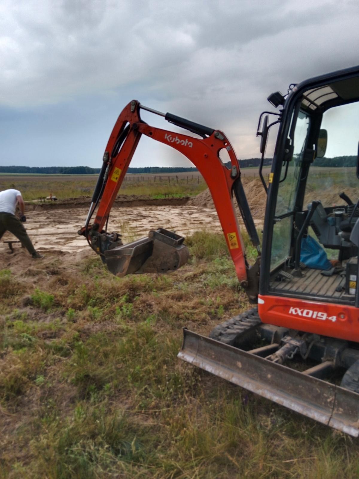 Pomarańczowa mini koparka Kubota KX019-4 w trakcie prac ziemnych na zewnątrz, z operatorem w tle i wykopem w oddali. Pochmurne niebo.