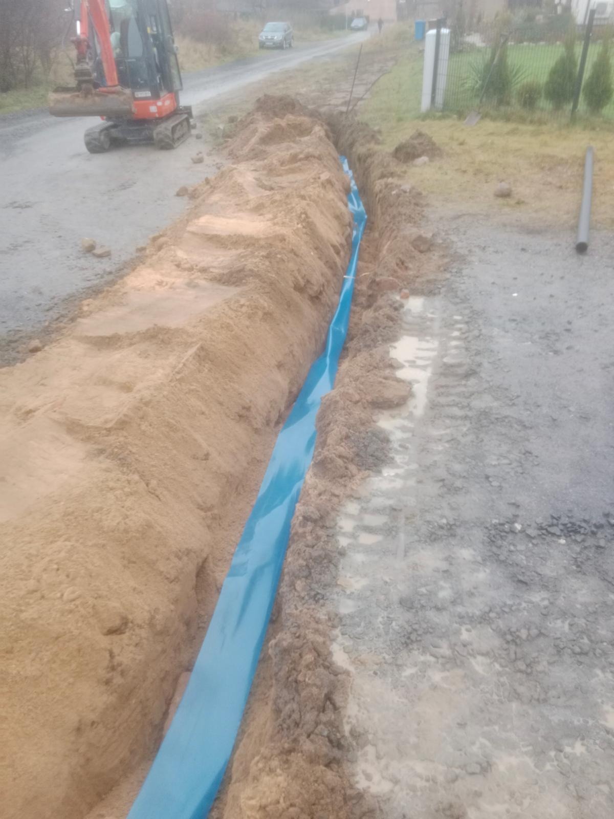 Wykop z niebieską folią ochronną, wykonany wzdłuż drogi. W tle widoczny mały czerwony koparka i samochód osobowy. Na ziemi ślady opon.