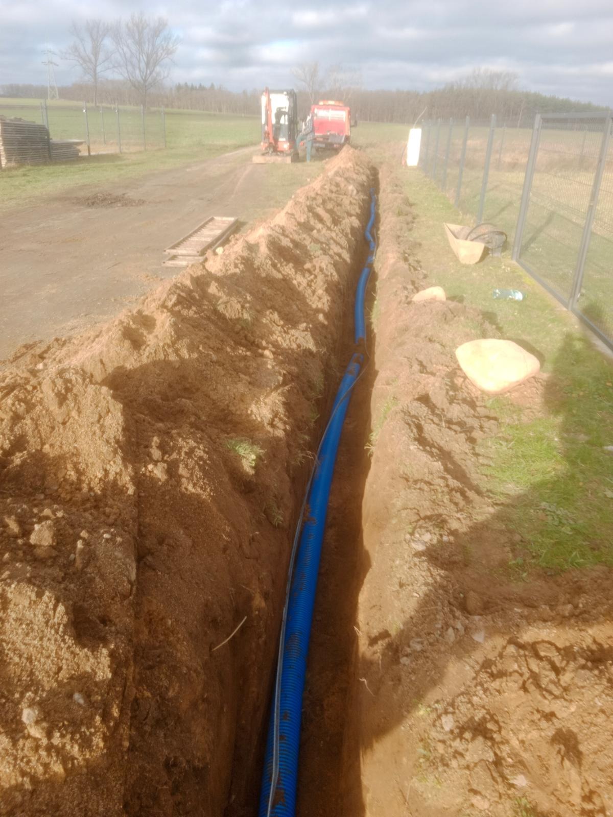 Wykop z niebieską rurą w ziemi, teren ogrodzony siatką, Polkowice. W tle maszyny budowlane i drzewa. Wykop pod instalacje.