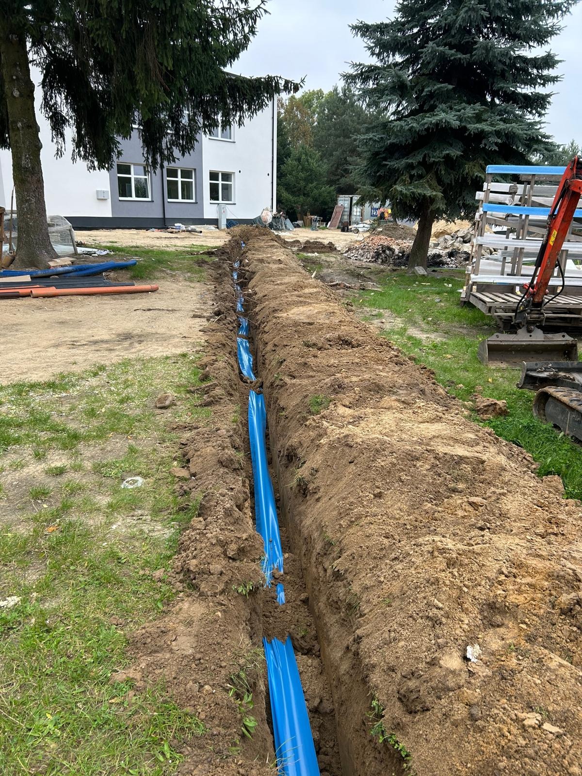 Długi wykop z niebieskimi rurami w ziemi, widoczny mały czerwony koparka i budynek w tle. Instalacja rur w wykopie.