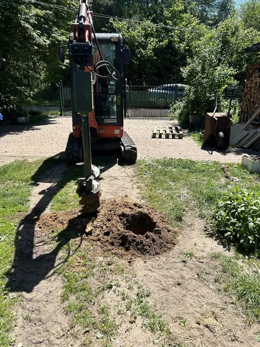 Wiercenie otworu w ziemi wiertnicą hydrauliczną zamontowaną na minikoparce. Widoczny wykopany otwór i cień wiertnicy. W tle trawa, drzewa i elementy zabudowy.