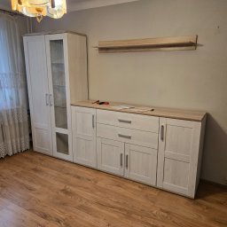 EDR - Jasny pokój po montażu mebli: szafa z witryną, komoda z szufladami i półka wisząca. Na blacie komody narzędzia i instrukcje. Drewniana podłoga i firanka w oknie.