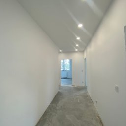 EDR - Wnętrze korytarza po szpachlowaniu i malowaniu ścian na biało. Widoczne nowe oświetlenie sufitowe i surowa podłoga przed wykończeniem.