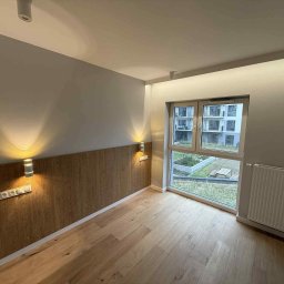 EDR - Pokój z drewnianą boazerią i parkietem, oświetlony kinkietami, z widokiem na zieleń za oknem. Minimalistyczny design, ciepłe światło, nowoczesne wykończenie.