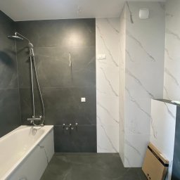 EDR - Wykończona łazienka z wanną, czarnymi i białymi płytkami imitującymi marmur, oraz kartonem po armaturze. Widok na baterię prysznicową i fragment toalety.