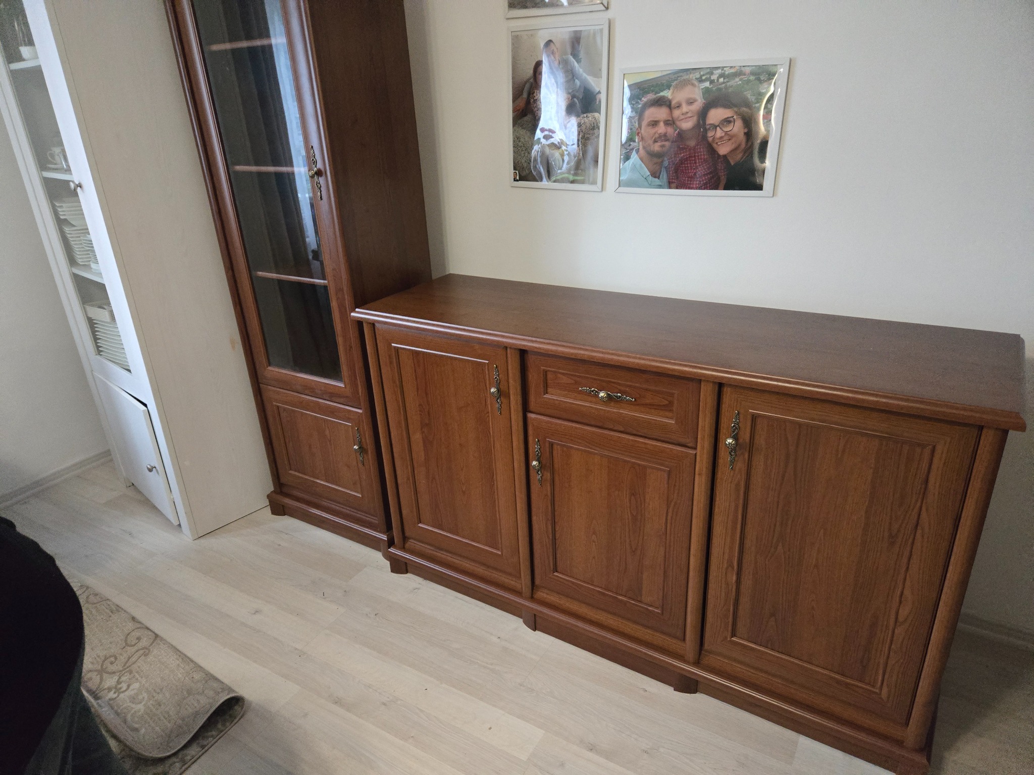 Zestaw mebli w salonie: ciemnobrązowa komoda z szafkami i szufladą, obok biały regał i witryna z ciemnego drewna. Na ścianie wiszą zdjęcia w ramkach.