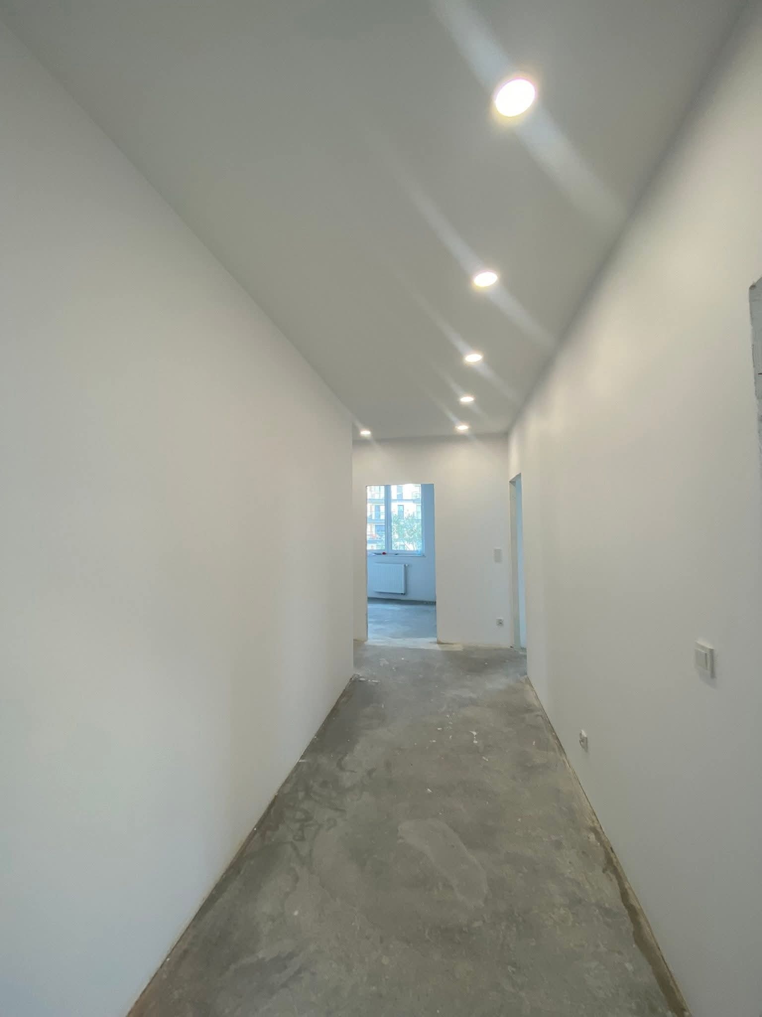 Wnętrze korytarza po szpachlowaniu i malowaniu ścian na biało. Widoczne nowe oświetlenie sufitowe i surowa podłoga przed wykończeniem.