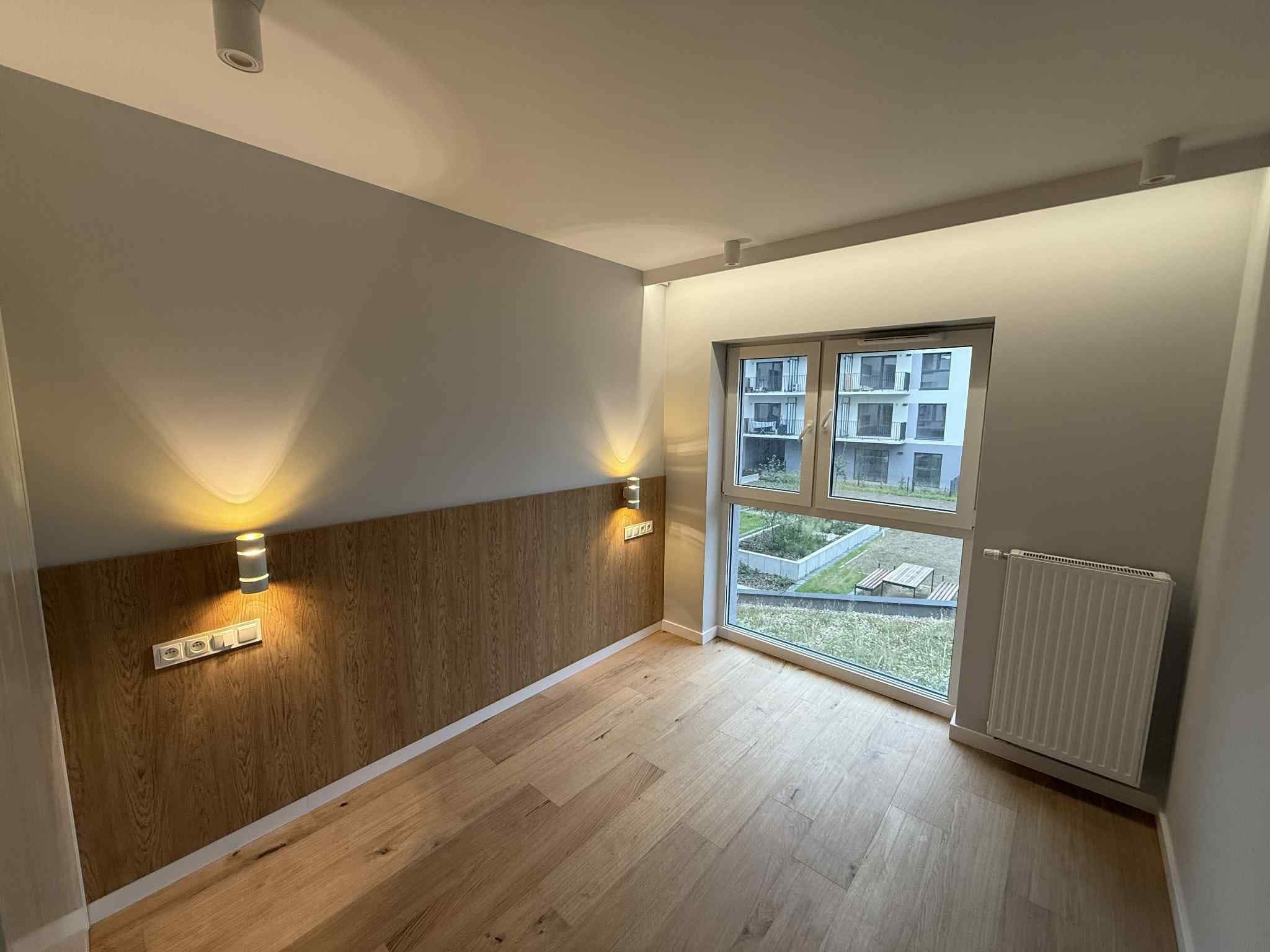 Pokój z drewnianą boazerią i parkietem, oświetlony kinkietami, z widokiem na zieleń za oknem. Minimalistyczny design, ciepłe światło, nowoczesne wykończenie.