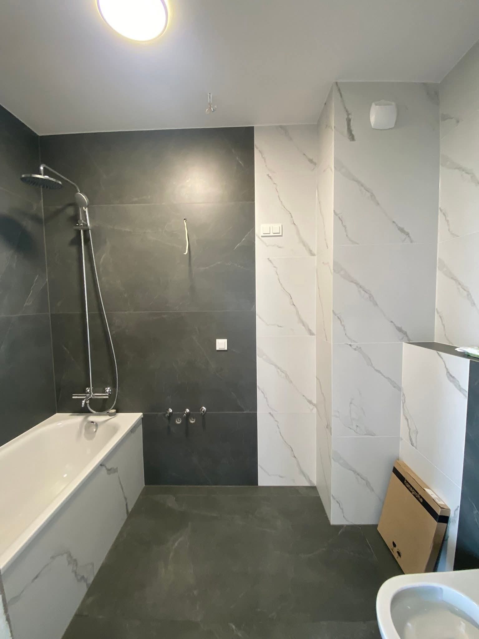 Wykończona łazienka z wanną, czarnymi i białymi płytkami imitującymi marmur, oraz kartonem po armaturze. Widok na baterię prysznicową i fragment toalety.