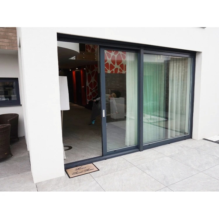 Nowoczesne, przesuwne drzwi balkonowe w grafitowej ramie, z widokiem na wnętrze domu i wyjście na taras z jasną, kamienną posadzką. Widoczna wycieraczka.