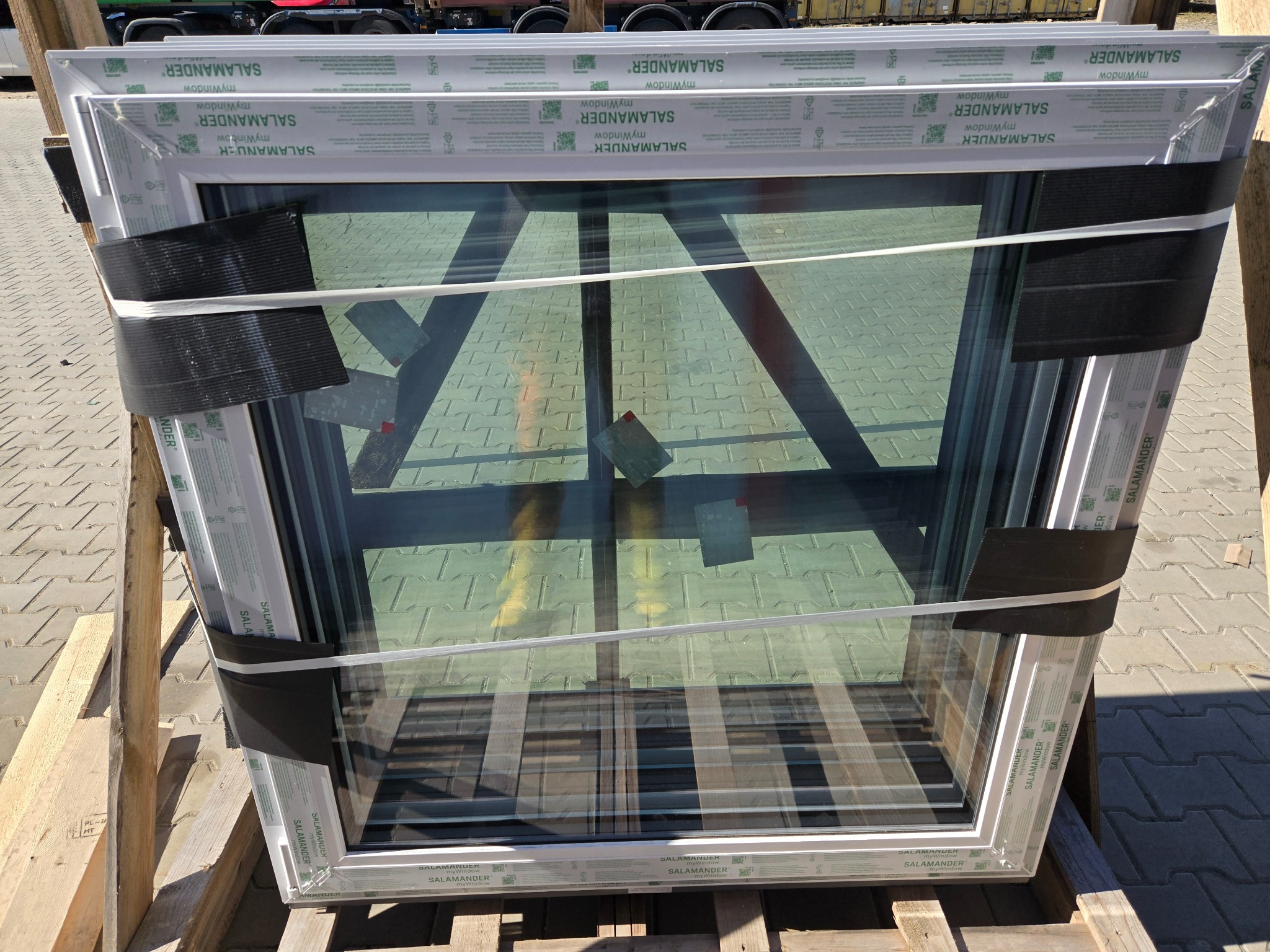 Nowe okno PCV marki Salamander, zabezpieczone taśmą i folią, gotowe do montażu, leży na drewnianej palecie na zewnątrz. Widoczne detale ramy i szyby.