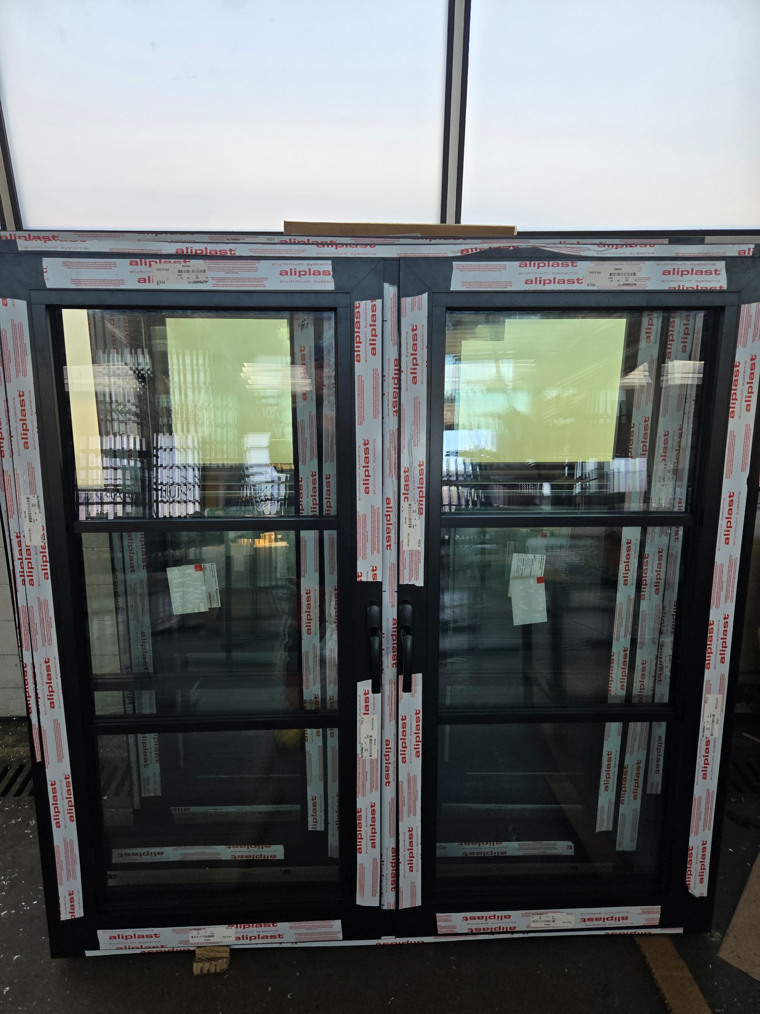 Nowe, dwuskrzydłowe okno z czarną ramą, zabezpieczone folią ochronną z nadrukiem 'aliplast'. Widoczne refleksy światła na szybach.